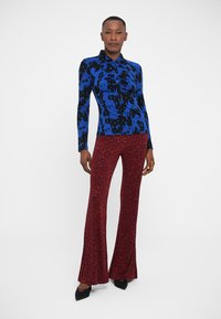 Langärmliges blaues Oberteil mit schwarz-abstraktem Muster; rote, ausgestellte Hose mit schwarzem Leopardenmuster. Die Modelle trägt schwarze High Heels. Schlicher weißer Hintergrund.