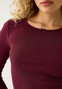 Bordowa, prążkowana bluzka z długim rękawem i okrągłym dekoltem. Posiada małą ozdobną kokardkę przy kołnierzyku. Miękka faktura i dopasowany krój.