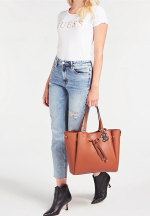 Femme portant un T-shirt blanc « Guess », un jean clair déchiré, des bottines noires, tenant un grand sac à main marron avec un pendentif logo en métal.