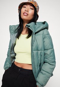 Groene pufferjack met een capuchon, voorzien van een doorgestikt ontwerp. Draagt over een lichtgele cropped top en is gecombineerd met zwarte broeken.