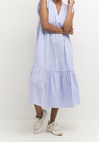 En person står med armarna över korsade, klädd i en blå och vit vertikalt randig midi-klänning och vita klumpiga sneakers framför en enkel bakgrund.