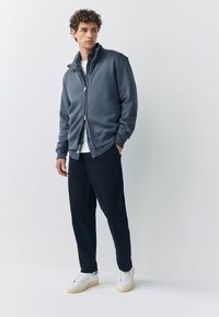 Grijze ritsvest met hoge kraag, gestreepte binnenvoering, gecombineerd met donkere broek en witte sneakers. Soepele stof, moderne snit.