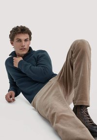 Marineblauwe geribbelde trui met een kwart rits, gecombineerd met beige corduroy broek en bruine veterschoenen. Model zit met een neutrale achtergrond.
