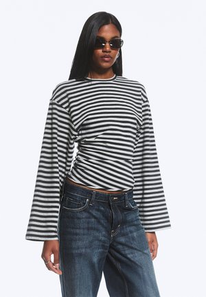 Frau mit langen, glatten Haaren, die schwarze Sonnenbrillen, ein schwarz-weiß gestreiftes Langarmshirt und dunkelblaue Jeans trägt und vor einem einfarbigen Hintergrund steht.