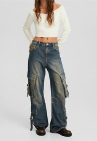 Biały prążkowany sweter z odkrytymi ramionami, krótki, zestawiony z niebieskimi dżinsowymi spodniami cargo, które mają duże kieszenie, suwaki i luźny krój.