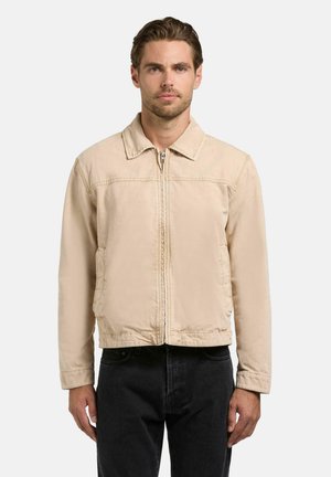 Mężczyzna o krótkich brązowych włosach, ubrany w beżową kurtkę na zamek i czarne spodnie, stojący na tle jednolitego jasnego tła.