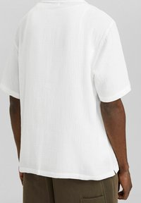 Persona dalla pelle scura che indossa una camicia bianca a maniche corte, ampia e dalla texture evidente, e pantaloni verde oliva, vista da dietro.