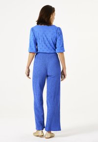 Haut texturé bleu avec des manches bouffantes, associé à un pantalon large assorti, tous deux en tissu léger. Des chaussures plates dorées complètent la tenue.