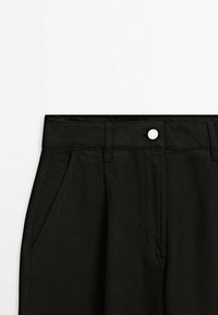 Pantalon noir taille haute avec fermeture à boutons, passants pour ceinture, poches latérales et détail plissé à l'avant sur fond blanc.
