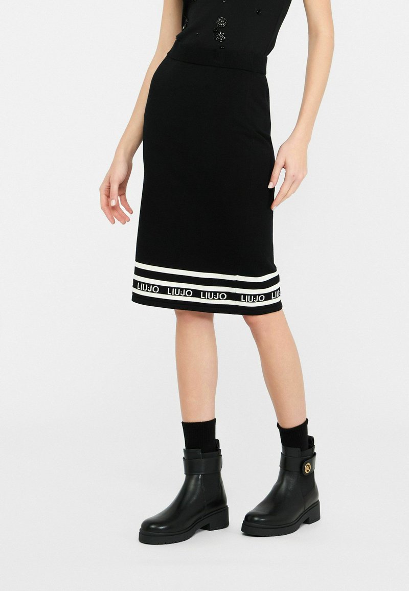 Jupe en jersey noir avec des accents à rayures blanches et une bande de logo à l'ourlet. Associée à des bottines noires et des chaussettes montantes. Texture lisse.