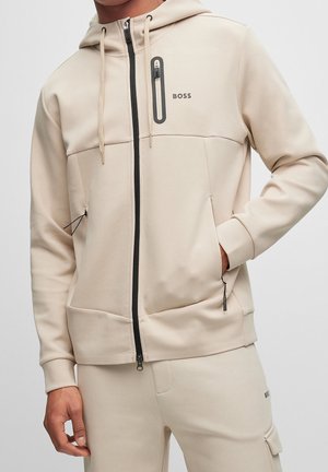Homme portant un sweat à capuche beige zippé avec cordons, fermeture éclair noire, poches poitrine et latérales, et pantalon beige assorti avec rabat sur la poche latérale.