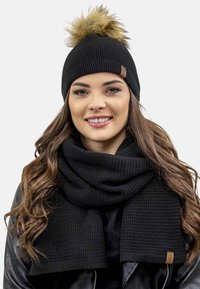 Zwarte gebreide beanie met een nep bont pompon en een bijpassende zwarte sjaal met een gestructureerd patroon. Beide accessoires zijn voorzien van een leren logo-label.