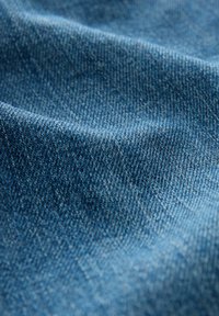 Denimtyg i en medelblå nyans, som visar textur med synliga vävningsmönster. Mjuk, lätt skrynklig yta, typisk för bomullsblandningar.