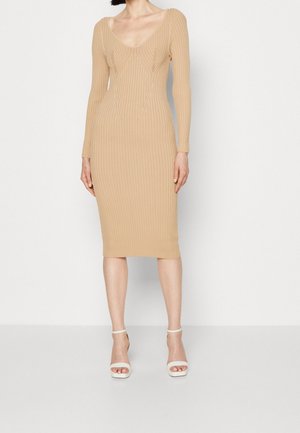 Kobieta w dopasowanej beżowej prążkowanej dzianinowej sukience midi z długimi rękawami i okrągłym dekoltem, w połączeniu z białymi sandałami na obcasie z odkrytymi palcami i paskiem wokół kostki.
