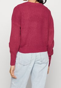 Textured, burgundy knit sweater with a round neck and ribbed cuffs, featuring a cropped design, worn over light denim jeans.