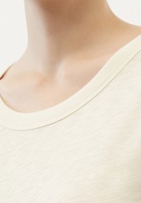 Cremefarbenes Baumwoll-T-Shirt mit weitem Ausschnitt, weicher Textur und leicht meliertem Muster auf dem Stoff.