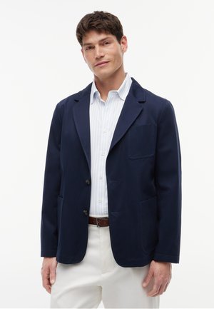 Mann trägt einen marineblauen Blazer über einem hellblau gestreiften Hemd und weißen Hosen, steht vor einem einfarbigen weißen Hintergrund.