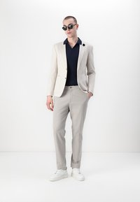 Lys beige blazer med gullknapper, marineblå ribbestrikket skjorte, lysegrå bukser og hvite joggesko. Solbriller gir kontrast til antrekket.