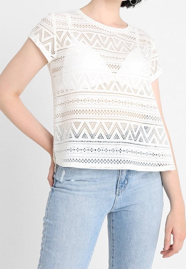 Personne portant un haut blanc à manches courtes avec un motif en dentelle et un jean bleu clair, une main reposée sur la hanche.