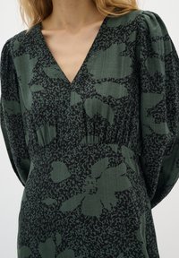 Grön blommig klänning med djupt V-halsringning, puffärmar och textil med struktur. Har svarta blommönster mot en grön bakgrund.