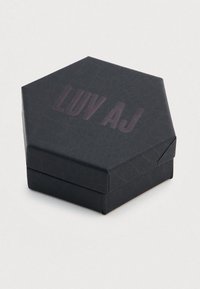 Εξάγωνο μαύρο κουτί με υφή στην επιφάνειά του. Το καπάκι φέρει ανάγλυφο το κείμενο "LUV AJ" σε σκούρο μωβ χρώμα. Κλείνει με ένα απλό δίπλωμα.