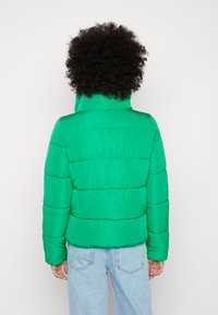 Grønn pufferjakke med høy krage, quiltet mønster og lange ermer. Kombinert med lyseblå jeans. Glatt tekstur, lett materiale.