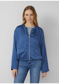 Kobieta ubrana w niebieską kurtkę z zamkiem błyskawicznym, pod którą ma pasiastą koszulę i niebieskie dżinsy, stoi na jednolitym szarym tle.