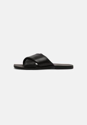 Sort flad sandal med brede krydsede læderremme og firkantet tådesign på en ensfarvet lys baggrund.