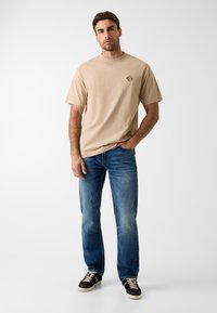 Beżowa bawełniana koszulka z krótkim rękawem i małym haftowanym logo, zestawiona z niebieskimi dżinsami o prostych nogawkach i czarnymi trampkami.
