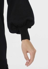 Svart stickad tröja med voluminösa puffärmar och ribbade manschetter. Tyget verkar texturerat och visar en åtsittande passform vid handleden.