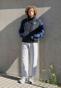 Sort letvægtsjakke med blå accenter, afslappede lysegrå bukser og grå sneakers; stående mod en betonvæg.