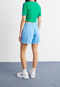 Zelený žebrovaný crop top s modrými akcenty, kombinovaný se světle modrými šortkami z látky s pružným pasem, obutý s bílými teniskami Adidas.