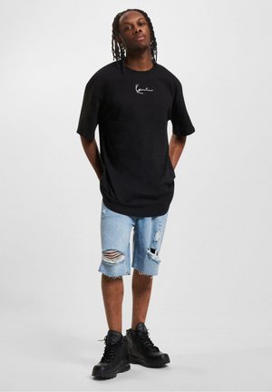 Čierne oversize tričko s bielym logom, doplnené svetlými denimbovými šortkami s otrhanými okrajmi a roztrhnutými detailmi, a čiernymi vysokými teniskami.