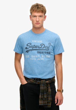 Blue short-sleeve t-shirt with a textured finish, featuring a vintage graphic print in dark blue and white. Black pants and plaid shirt tied at the waist.