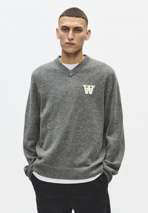 Junger Mann mit kurzen Haaren, der einen grauen V-Ausschnitt-Pullover mit einem weißen "W"-Aufnäher und schwarzen Hosen trägt, steht mit den Händen in den Taschen.