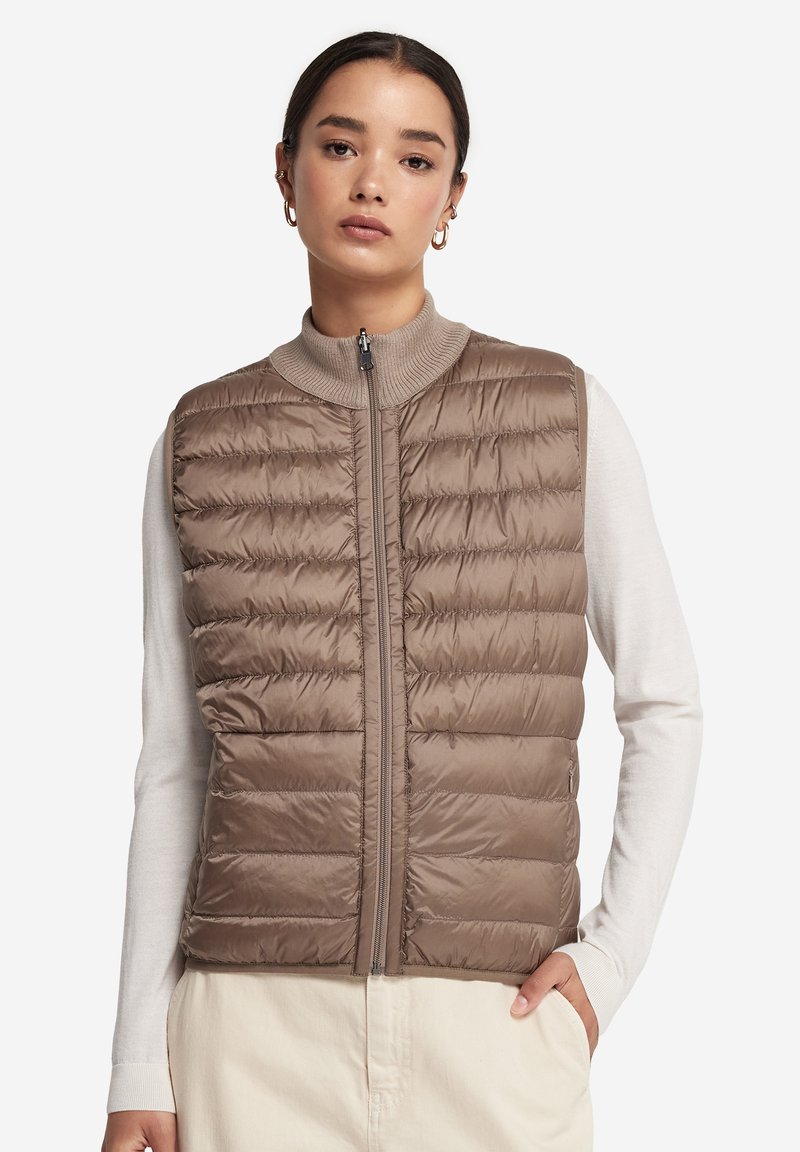 Frau trägt eine taupefarbene, gesteppte ärmellose Weste über einem weißen Langarmshirt und cremefarbenen Hosen, steht mit einer Hand in der Tasche.