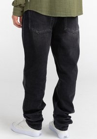 Persona vestida con pantalones vaqueros negros de corte holgado, zapatillas blancas y una camisa de manga larga verde olivo, de pie contra un fondo liso.