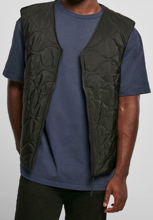Homme portant un gilet matelassé sans manches noir sur un t-shirt bleu foncé à manches courtes, avec le gilet dézippé devant.