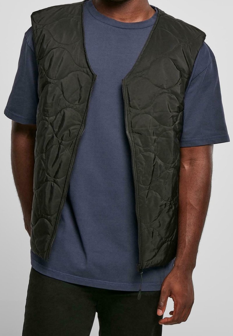 Homme portant un gilet matelassé sans manches noir sur un t-shirt bleu foncé à manches courtes, avec le gilet dézippé devant.