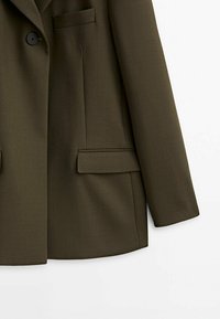 Blazer vert olive avec un bouton noir, deux poches à rabat et une poche passepoilée sur la poitrine, présenté sur fond blanc.