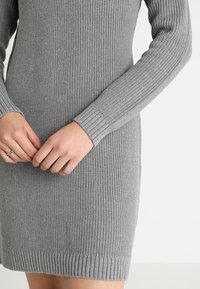 Graues Strickkleid mit langen Ärmeln, das über eine gerippte Textur, eine figurbetonte Passform und einen Saum verfügt. Die Hände sind auf der Taille positioniert.