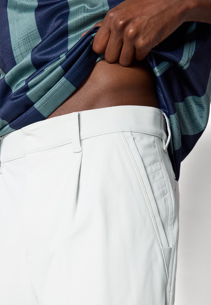 Pantaloni bianchi sartoriali con pieghe frontali, tasche laterali e una texture liscia, abbinati a una camicia a righe blu e verde.