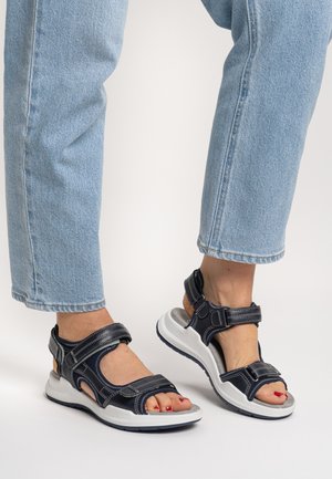 Navy blue sandals with adjustable straps, textured synthetic material, and a white cushioned sole. Features open-toe design and contrasting stitching.