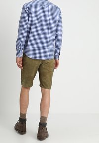 Mann steht mit dem Rücken zur Kamera, trägt ein blau kariertes Hemd, olivgrüne Shorts, braune Socken und dunkelbraune Stiefel vor einem schlichten Hintergrund.
