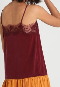 Top senza maniche bordeaux con rifiniture in pizzo attorno al colletto, tessuto morbido e delicate frange lungo i bordi.