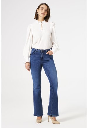 Vrouw die een witte blouse met lange mouwen, blauwe hooggetailleerde wijde jeans en beige hoge hakken draagt, staand tegen een effen achtergrond.