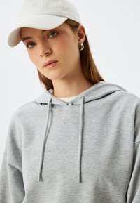 Grijze hoodie met een gestructureerde uitstraling, voorzien van een trekkoord en een gelaagde kraag. Een witte pet completeert de outfit.
