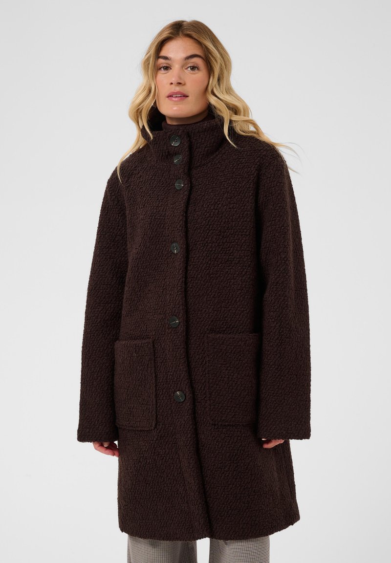 Femme portant un long manteau texturé marron foncé avec de gros boutons et deux poches avant, debout devant un fond uni clair.