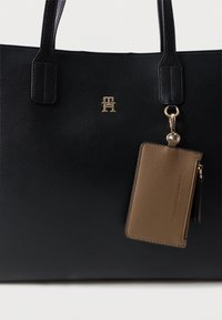 Czarna torebka tote z fakturowanej skóry, z podwójnymi uchwytami u góry i złotym logo. W zestawie znajduje się odpinany brązowy portfel na karty z zamkiem.