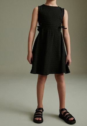 Schwarzes ärmelloses Kleid aus strukturiertem Stoff, in der Taille mit Bändern gerafft. Trägt man mit schwarzen Riemchensandalen.